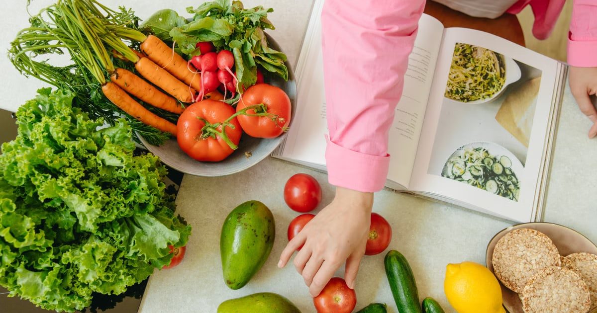 Top view of fresh vegetables and hands arranging them with a recipe book, promoting healthy eating.