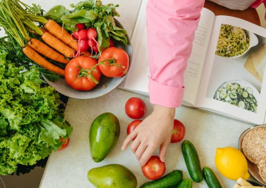 Top view of fresh vegetables and hands arranging them with a recipe book, promoting healthy eating.