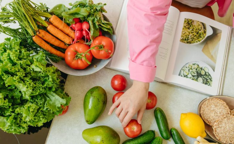 Top view of fresh vegetables and hands arranging them with a recipe book, promoting healthy eating.