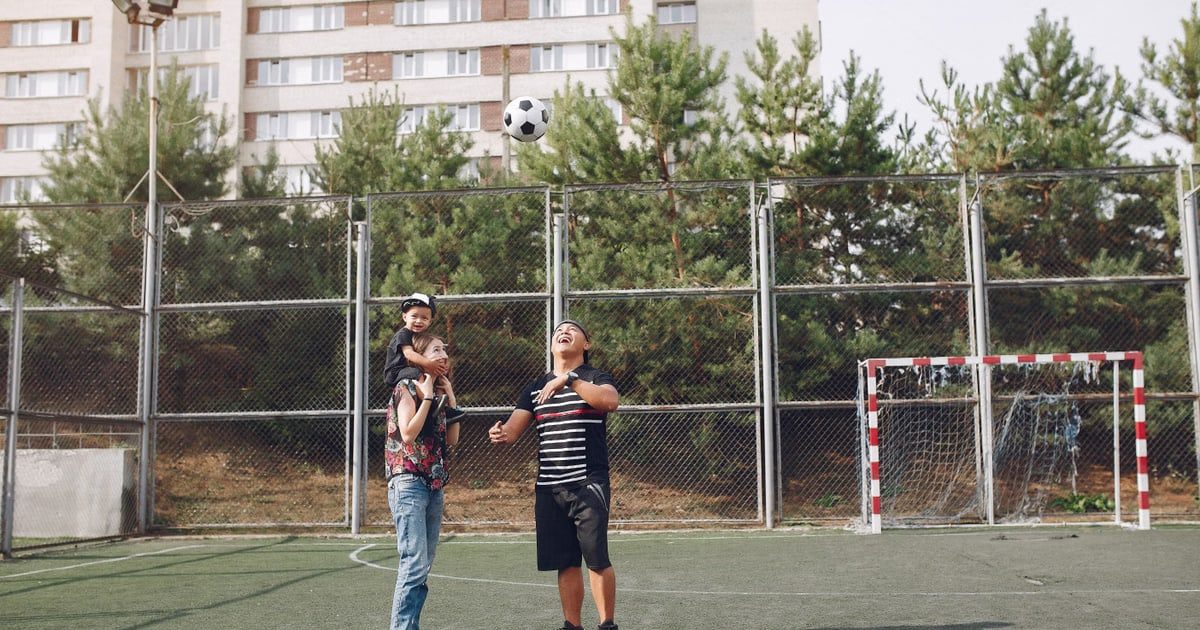 Full body cheerful father playing with football ball and mother giving piggyback ride to cute son while walking together on sports ground