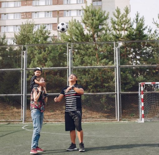 Full body cheerful father playing with football ball and mother giving piggyback ride to cute son while walking together on sports ground