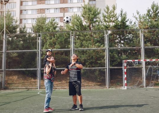 Full body cheerful father playing with football ball and mother giving piggyback ride to cute son while walking together on sports ground