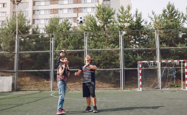 Full body cheerful father playing with football ball and mother giving piggyback ride to cute son while walking together on sports ground