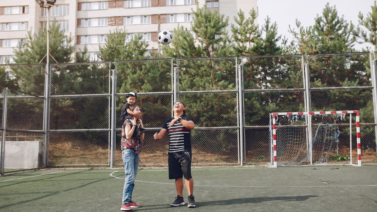 Full body cheerful father playing with football ball and mother giving piggyback ride to cute son while walking together on sports ground